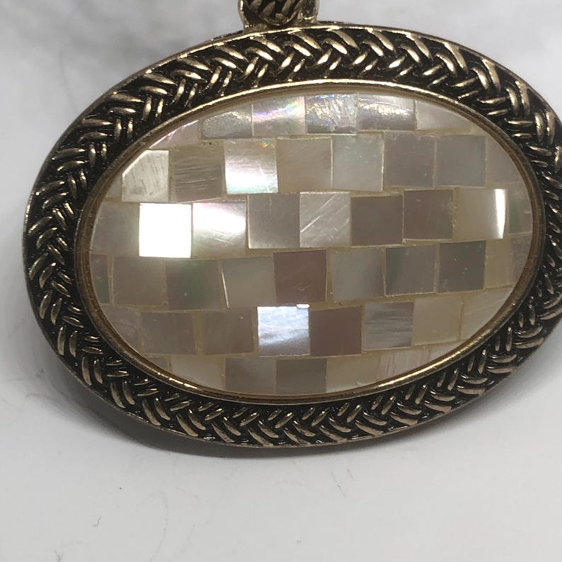 May include: A gold oval pendant with a braided border and a center made of white mother of pearl tiles.