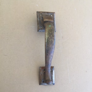 May include: A vintage, rusty metal door handle with a curved design. The handle is attached to two square plates with visible screw holes.