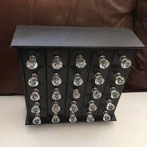 May include: A black wooden drawer cabinet with 24 clear glass knobs. The cabinet is a small size and has a rectangular shape.