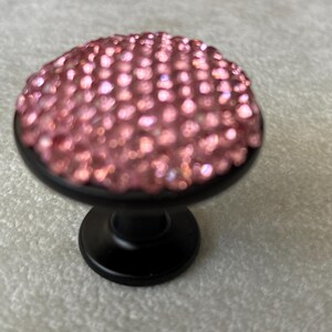 May include: A round cabinet knob featuring a matte black metal base and a top surface densely covered in small, sparkling pink crystals. The knob has a circular, flat-topped design with a short, tapered stem that connects to a circular base plate.