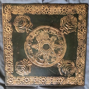 May include: A square, dark green tile with a raised, gold-colored design. The design features four stylized roses and a central circular motif with intricate floral patterns.
