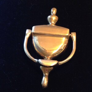 May include: A gold-toned door knocker in the shape of a traditional urn with a rounded top and a decorative ring.