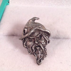 May include: A silver pewter pin of a wizard with a pointed hat and a long beard. The wizard has a stern expression on his face.