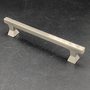 May include: A brushed nickel cabinet pull with a rectangular bar and square ends. The pull is about 6 inches long.