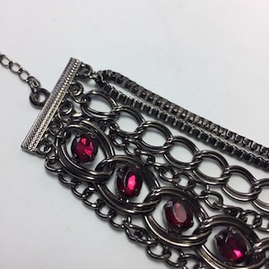 May include: A multi-strand bracelet with a gunmetal finish and red rhinestone accents. The bracelet features a variety of chain styles, including box chain, cable chain, and curb chain.