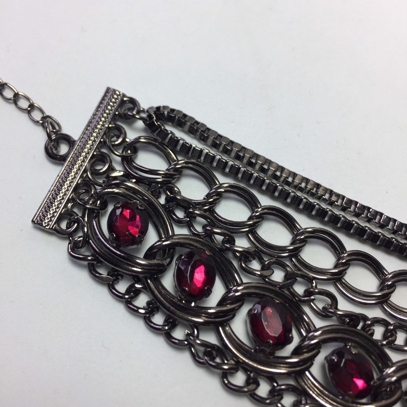May include: A multi-strand bracelet with a gunmetal finish and red rhinestone accents. The bracelet features a variety of chain styles, including box chain, cable chain, and curb chain.