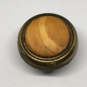 May include: A round, brass knob with a wood inlay. The wood has a light brown color with a darker brown swirl pattern.