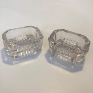 May include: Two clear glass salt cellars with a ribbed design and three legs each.