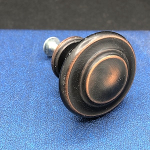 May include: A round, black cabinet knob with a copper finish. The knob has a raised, circular design.