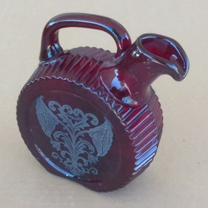 Puede incluir: Una pequeña jarra redonda de vidrio rojo con un diseño grabado en plata de un motivo floral. La jarra tiene un asa y un pico.