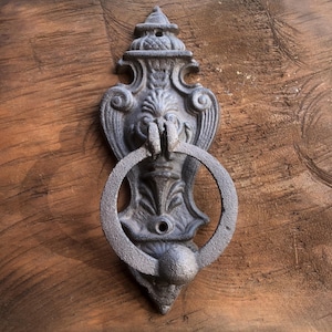 May include: A detailed, antique-style door knocker made of dark gray metal. The knocker features an ornate design with a circular handle and decorative elements. It is mounted on a wooden door with visible wood grain.