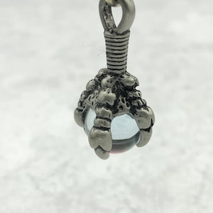May include: A silver pendant with a clear glass orb and a detailed claw design. The orb has a hint of pink in the center.