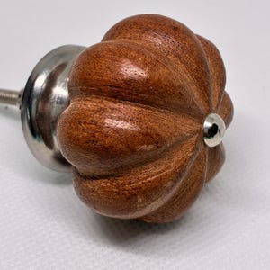 May include: A brown wooden cabinet knob with a rounded, pumpkin-like shape and vertical ridges. The knob features a smooth, polished finish with visible wood grain patterns and a small, shiny silver-toned metal center cap and base.