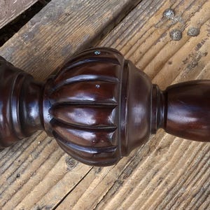 May include: A dark brown, wooden furniture leg or finial with a bulbous, ribbed center and a rounded end. The piece has a polished finish and a threaded bolt extending from one end. It rests on a wooden surface.