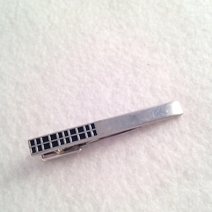 May include: A silver tie bar with a black geometric design. The tie bar is about 2 inches long.