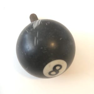 以下が含まれることがあります： 白い円の中に数字の8が入った黒いビリヤードボール。ねじ付きの金属棒が付いています。ボールの表面には白い傷がいくつかあります。