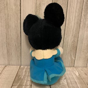 May include: A plush toy featuring a black head with large ears and a blue dress. The toy has a white collar and a light blue trim on the dress. The toy is positioned against a light wood-grain background.