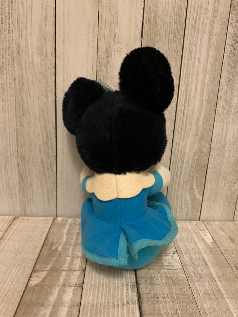 May include: A plush toy featuring a black head with large ears and a blue dress. The toy has a white collar and a light blue trim on the dress. The toy is positioned against a light wood-grain background.
