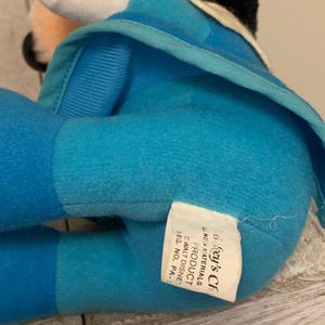 May include: A plush toy in shades of blue, with a white and black head. The toy has a tag that reads "Gey's CF PRODUCT" and "WALT DISNEY". The toy is made of soft material.