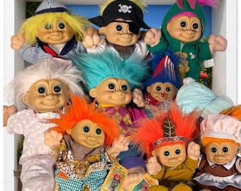 May include: A collection of ten troll dolls with various hair colors and outfits, including a pirate with an eye patch, a sailor, a green hooded figure, and others in patterned clothing, all displayed together in a white square frame.
