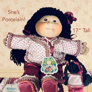 May include: A vintage porcelain doll, 17 inches tall, with black yarn hair and a burgundy bow. The doll wears a floral print dress with burgundy trim and matching pants. A smaller doll is also present.