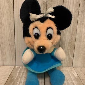 May include: A plush Minnie Mouse doll with black ears and a light blue dress. The doll has a beige face, a black nose, and a white bow. The doll's arms and legs are light blue.