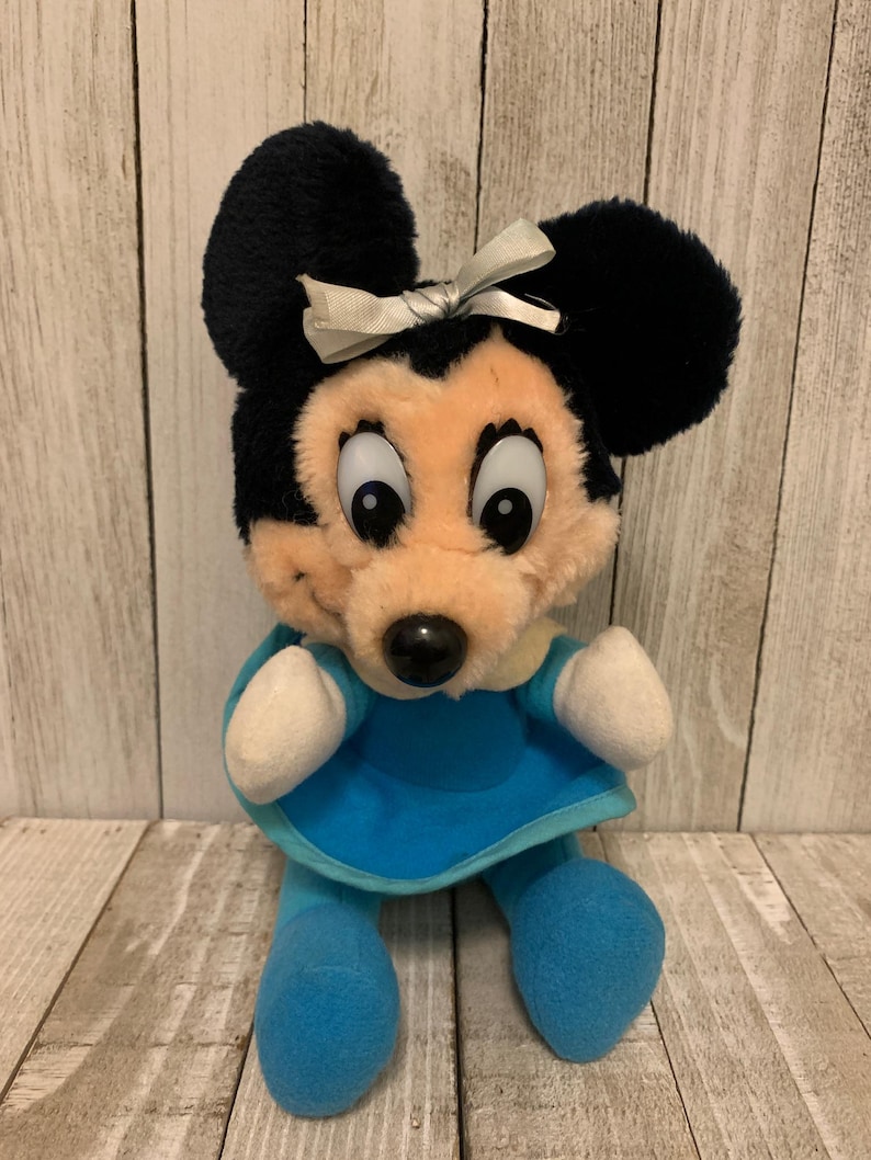 May include: A plush Minnie Mouse doll with black ears and a light blue dress. The doll has a beige face, a black nose, and a white bow. The doll's arms and legs are light blue.