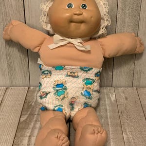 May include: A vintage Cabbage Patch Kid doll with a soft, peach-colored body and a bald head. The doll wears a white bonnet with lace trim and a diaper with a repeating pattern of teddy bears. A white bow is tied at the neck.