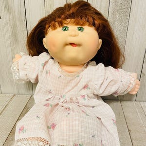 May include: A vintage doll with auburn hair and green eyes, wearing a light pink dress with a floral pattern and lace trim. The doll's dress is tied with a bow at the waist. The doll is a classic toy.