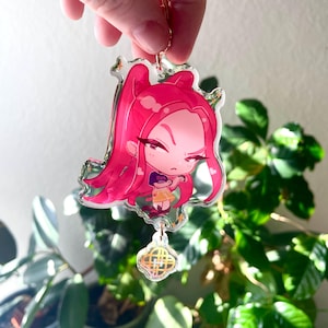 Op de afbeelding: Een heldere acryl bedel met een rozeharige anime-stijl karakter met een boze uitdrukking. De bedel heeft een goudkleurige ketting en een decoratieve hanger aan de onderkant. Het karakter draagt een gele jurk en een paarse top.
