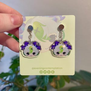 May include: A pair of silver earrings with a clear acrylic charm featuring a cartoon character with green hair, purple eyes, and a green cross on their forehead. The character is surrounded by a purple and green halo. The earrings are on a white background with the text "@eveningcontemplation" and social media icons.