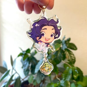 Op de afbeelding: Een heldere acryl sleutelhanger met een stripfiguur met paars haar en een witte outfit. De sleutelhanger heeft een goudkleurige ring en een bedel met een goud en iriserend ontwerp. Het personage lacht en rent.