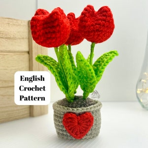Puede incluir: Tulipanes rojos tejidos a crochet en una maceta gris con un corazón rojo. La maceta está sobre una superficie de madera. Patrón de crochet en inglés.
