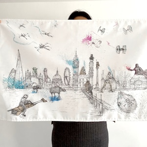 Op de afbeelding: Een witte canvas draagtas met een zwart-witte illustratie van Londense bezienswaardigheden en Star Wars-personages. De illustratie omvat Big Ben, het London Eye en de Millennium Bridge. Star Wars-personages zijn Darth Vader, Yoda en R2-D2.
