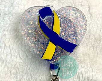 May include: A clear heart-shaped badge holder with a blue and yellow ribbon inside. The holder is filled with iridescent glitter and has a silver swivel clip and a blue glitter button.