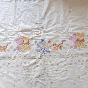 May include: White Winnie the Pooh themed bedding with a blue and white border. The bedding features a repeating pattern of stars, flowers, and hearts. The bottom of the bedding features a row of Winnie the Pooh characters, including Pooh Bear, Piglet, Eeyore, and Tigger.