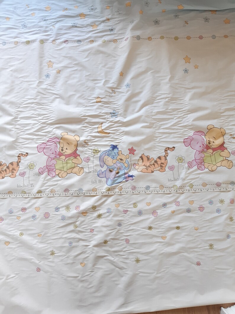 May include: White Winnie the Pooh themed bedding with a blue and white border. The bedding features a repeating pattern of stars, flowers, and hearts. The bottom of the bedding features a row of Winnie the Pooh characters, including Pooh Bear, Piglet, Eeyore, and Tigger.