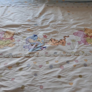 May include: White bedding with a colourful Winnie the Pooh and friends print. The print features Winnie the Pooh, Piglet, Tigger, Eeyore, and a blue character reading a book.