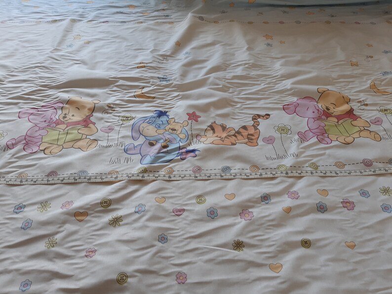 May include: White bedding with a colourful Winnie the Pooh and friends print. The print features Winnie the Pooh, Piglet, Tigger, Eeyore, and a blue character reading a book.
