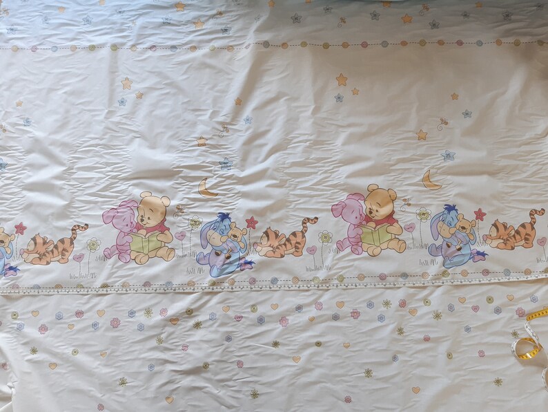 May include: White fabric featuring a repeating pattern of Winnie the Pooh and his friends. The characters are depicted in a cartoon style and are surrounded by stars and flowers. The fabric is suitable for crafting projects, such as quilts, blankets, and cushions.