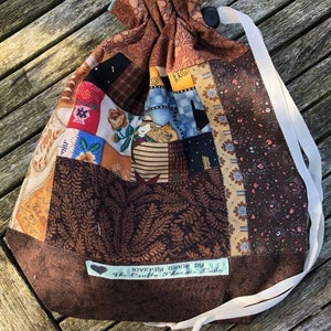 Op de afbeelding: Een bruine patchwork-trektassen met een wit trekkoord en een label met de tekst "lovingly made by The Crafty Phoenix Lady".