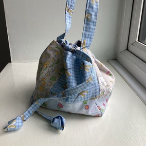 Puede incluir: Una pequeña bolsa de cordón azul y blanco con estampado floral y libélula. La bolsa tiene un solo asa y una parte superior cinchada.