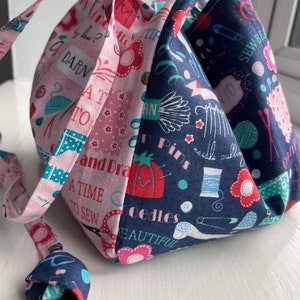 Puede incluir: Una bolsa de cordón de tela rosa y azul con temática de costura. La bolsa está decorada con varias imágenes y textos relacionados con la costura, incluyendo "DARN", "AT TO", "SEW BE IT!" y "A TIME TO SEW".