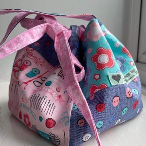 Puede incluir: Una bolsa de cordón de tela rosa y azul con un diseño de patchwork. La bolsa presenta una variedad de estampados con temática de costura, incluidos botones, agujas e hilo. La bolsa tiene un cordón rosa y una etiqueta que dice "Hecho por Rosie B."