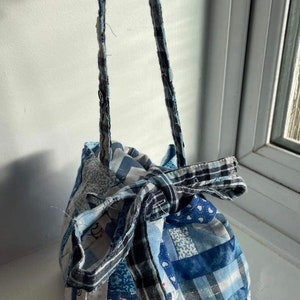 Könnte beinhalten: Eine kleine blaue und weiße Patchwork-Kordeltasche mit einem langen blauen und weißen Riemen. Die Tasche ist aus verschiedenen Stoffen mit verschiedenen Mustern gefertigt, darunter Karo, Streifen und Punkte.
