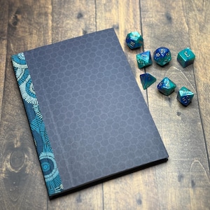 Op de afbeelding: Een donkerblauw notitieboek met een honingraatpatroon en een patroon turquoise rug. Verschillende turquoise en blauwe veelvlakkige dobbelstenen liggen verspreid rond het notitieboek op een houten oppervlak. De dobbelstenen hebben verschillende vormen en maten.