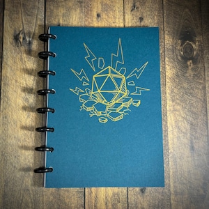 Pode incluir: Um caderno azul-petróleo com encadernação preta. A capa apresenta uma ilustração dourada de um dado de 20 lados, raios e rochas quebradas. O caderno está aberto sobre uma superfície de madeira.