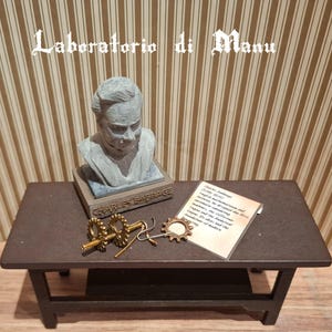 Può includere: Un tavolo marrone scuro in miniatura con un busto grigio di Charles Babbage, un foglio di carta con testo e ingranaggi in ottone. Il busto poggia su una base con la scritta "CHARLES BABBAGE". Il testo "Laboratorio di Manu" è nella parte superiore dell'immagine.