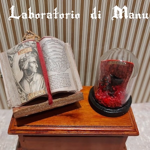 May include: A miniature wooden box with a book and a glass dome with a red object inside. The book is open and has a red bookmark. The book has the title "Inferno" on the cover. The box is on a wooden surface.