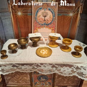 Puede incluir: Pequeños cálices, patenas y otros objetos religiosos dorados están dispuestos sobre una mesa blanca con encaje. Un letrero decorativo con imágenes religiosas está en el fondo. El texto "Laboratorio di Manu" está en la parte superior.